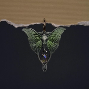 Puede incluir: Un colgante detallado con un diseño de polilla con alas verdes y cuerpo plateado. El colgante tiene un pequeño cristal iridiscente colgando de la parte inferior. La polilla está suspendida de un aro dorado.