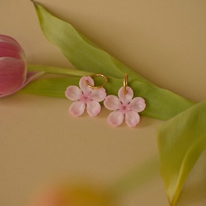 Puede incluir: Un par de pendientes rosas con forma de flor y aros dorados. Los pendientes están sobre un fondo beige suave con una hoja verde y un tulipán rosa. Los pendientes tienen un diseño delicado y femenino.