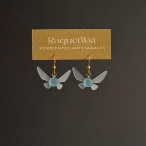 Puede incluir: Un par de pendientes con un diseño de hada azul claro y ganchos dorados. Los pendientes se muestran en una tarjeta que dice "RaquelWst PENDIENTES ARTESANALES".