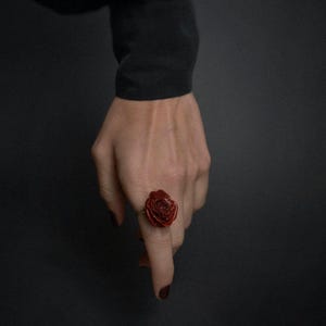 Op de afbeelding: Een rode rozenring met een gouden band wordt op een vinger getoond. De ring is het middelpunt, met de hand en een zwarte mouw op de achtergrond. Het rozenontwerp van de ring is gedetailleerd en gestructureerd.