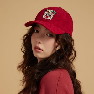 May include: A red corduroy baseball cap with a curved peak. The cap has an embroidered tiger face design with the text "Flavours" above it. The cap is worn by a person with long, wavy brown hair.