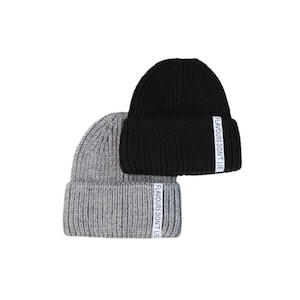 May include: Two knitted beanies, one grey and one black, are stacked. Each beanie has a white label with the text "FLAVOURS DON'T LIE" on the folded brim. The beanies are made of a thick, ribbed knit material.