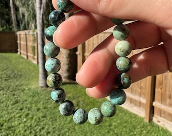 May include: A turquoise beaded bracelet with a mix of green and black tones. The round beads are strung together, creating a stretch bracelet. The bracelet is held up against a background of green grass and a wooden fence.