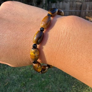 May include: A beaded bracelet featuring oval-shaped tiger's eye stones and small black beads. The bracelet is strung together, showcasing the natural patterns and colors of the tiger's eye stones.