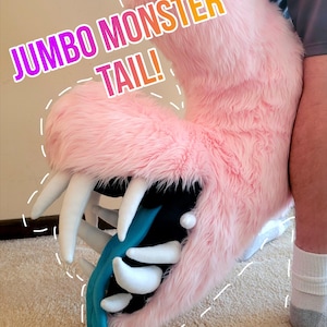 May include: A jumbo pink faux fur monster tail featuring a wide open mouth with white fabric teeth and a long teal tongue. The text JUMBO MONSTER TAIL! is visible above the tail, and PROBABLYASTERSUITS is printed at the bottom.