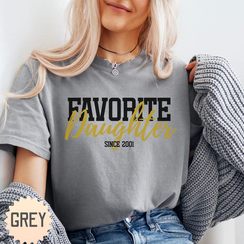 Puede incluir: Una camiseta gris con las palabras "FAVORITE DAUGHTER" en letras negras y doradas, con "SINCE 2001" debajo. La camiseta es de color gris liso. La palabra "GREY" est&aacute; en un peque&ntilde;o cuadro en la parte inferior izquierda.