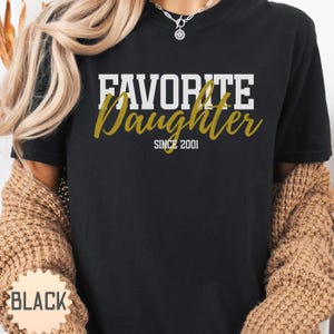 Puede incluir: Camiseta negra con la frase "FAVORITE Daughter" en letras blancas y doradas. El texto "SINCE 2001" est&aacute; debajo. La camiseta es de cuello redondo.