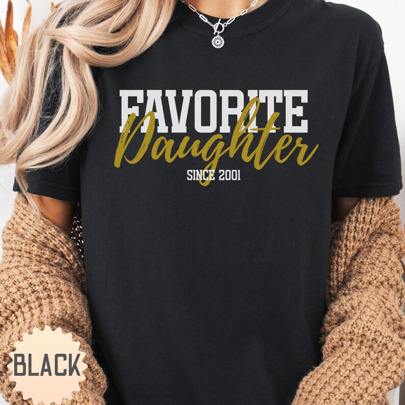 Puede incluir: Camiseta negra con la frase "FAVORITE Daughter" en letras blancas y doradas. El texto "SINCE 2001" est&aacute; debajo. La camiseta es de cuello redondo.