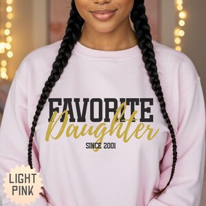 Puede incluir: Sudadera rosa claro con el texto "FAVORITE Daughter SINCE 2001" en letras negras y doradas. Las palabras "LIGHT PINK" est&aacute;n en forma de estrella en la esquina inferior izquierda. La sudadera es de cuello redondo.