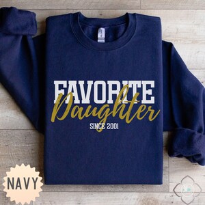 Puede incluir: Sudadera azul marino con las palabras "FAVORITE Daughter" en letras blancas y doradas. El texto "SINCE 2001" est&aacute; debajo de la palabra "Daughter". La sudadera tiene cuello redondo y mangas largas.