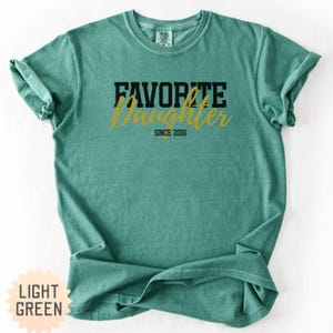 Puede incluir: Camiseta verde claro con las palabras "FAVORITE DAUGHTER SINCE 2001" impresas en negro y dorado. La camiseta tiene cuello redondo y mangas cortas. El texto "LIGHT GREEN" est&aacute; impreso sobre un fondo amarillo claro.