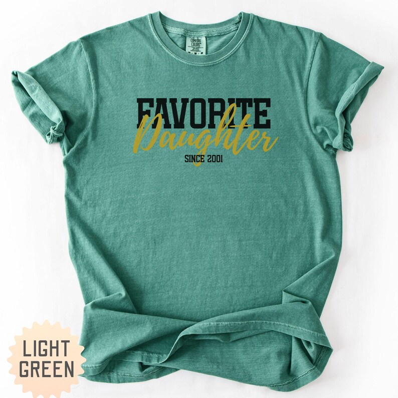 Puede incluir: Camiseta verde claro con las palabras "FAVORITE DAUGHTER SINCE 2001" impresas en negro y dorado. La camiseta tiene cuello redondo y mangas cortas. El texto "LIGHT GREEN" est&aacute; impreso sobre un fondo amarillo claro.