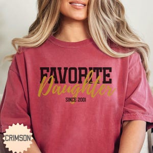 Puede incluir: Una camiseta granate con las palabras "FAVORITE Daughter" en letras negras y doradas. El texto "SINCE 2001" est&aacute; debajo de la palabra "Daughter". La camiseta tiene mangas cortas y cuello redondo.