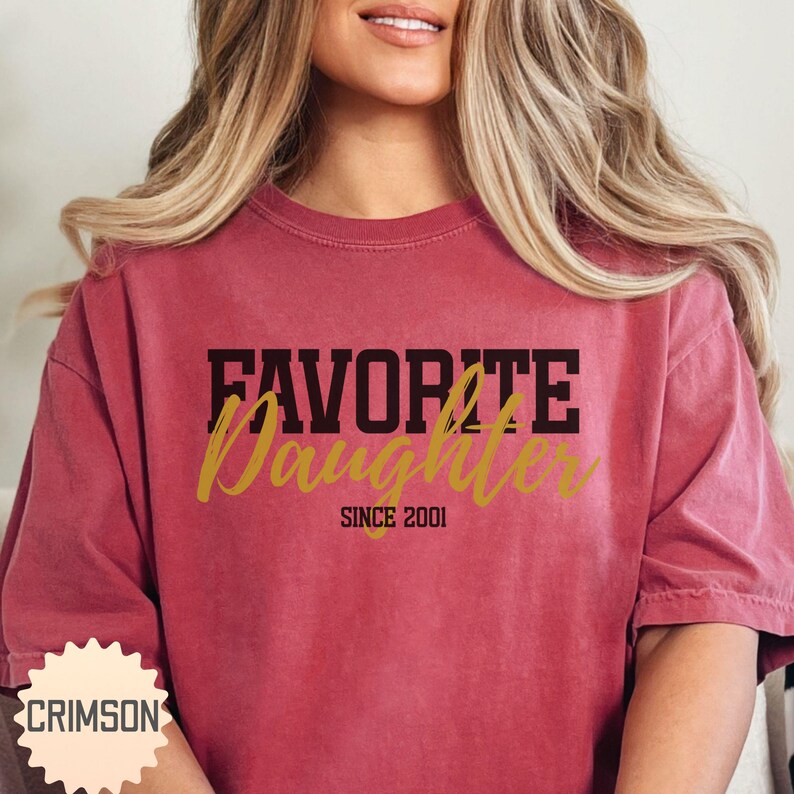 Puede incluir: Una camiseta granate con las palabras "FAVORITE Daughter" en letras negras y doradas. El texto "SINCE 2001" est&aacute; debajo de la palabra "Daughter". La camiseta tiene mangas cortas y cuello redondo.