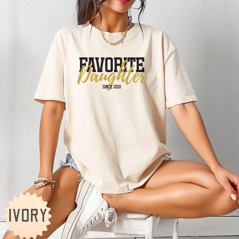 Puede incluir: Camiseta color marfil con el texto "FAVORITE Daughter" en letras negras y doradas, con "SINCE 2001" debajo. La camiseta tiene mangas cortas y un corte holgado.