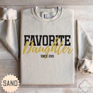 Puede incluir: Una sudadera de color arena con las palabras "FAVORITE Daughter" en letras negras y doradas. El texto "SINCE 2001" est&aacute; debajo. La sudadera tiene cuello redondo y mangas largas.