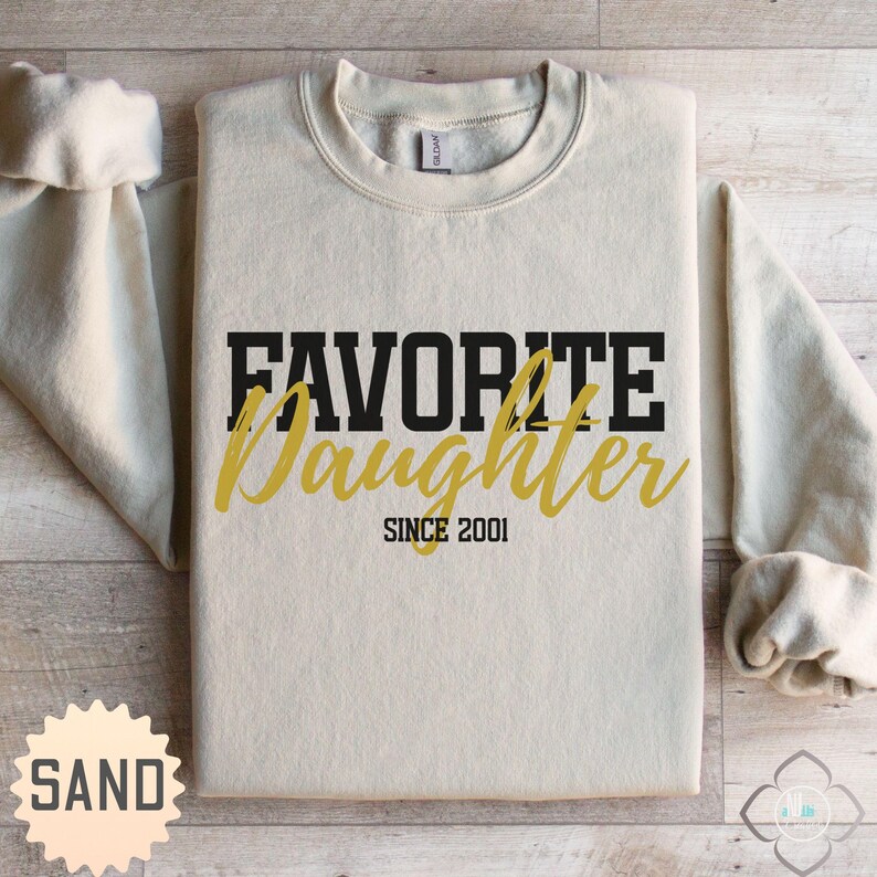 Puede incluir: Una sudadera de color arena con las palabras "FAVORITE Daughter" en letras negras y doradas. El texto "SINCE 2001" est&aacute; debajo. La sudadera tiene cuello redondo y mangas largas.