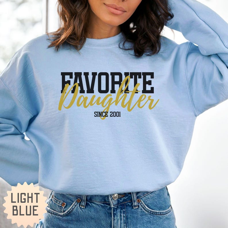 Puede incluir: Sudadera azul claro con el texto "FAVORITE Daughter SINCE 2001" en letras negras y doradas. La sudadera es de cuello redondo y est&aacute; hecha de un material suave. Las palabras "LIGHT BLUE" est&aacute;n impresas en un recuadro en la esquina inferior izquierda.