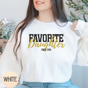 Puede incluir: Sudadera blanca con el texto "FAVORITE Daughter SINCE 2001" en letras negras y doradas. La sudadera es de color blanco liso y tiene mangas largas. La palabra "WHITE" est&aacute; impresa en la esquina inferior izquierda.