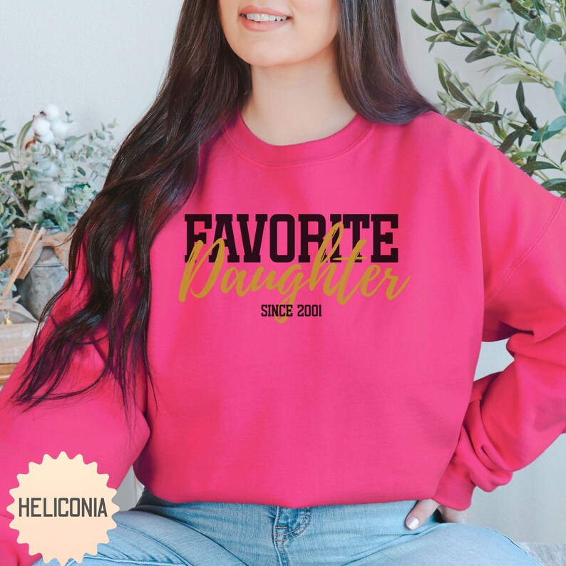 Puede incluir: Una sudadera magenta con el texto "FAVORITE Daughter" en letras negras y doradas. El texto "SINCE 2001" est&aacute; debajo de la palabra "Daughter". La sudadera es de cuello redondo.