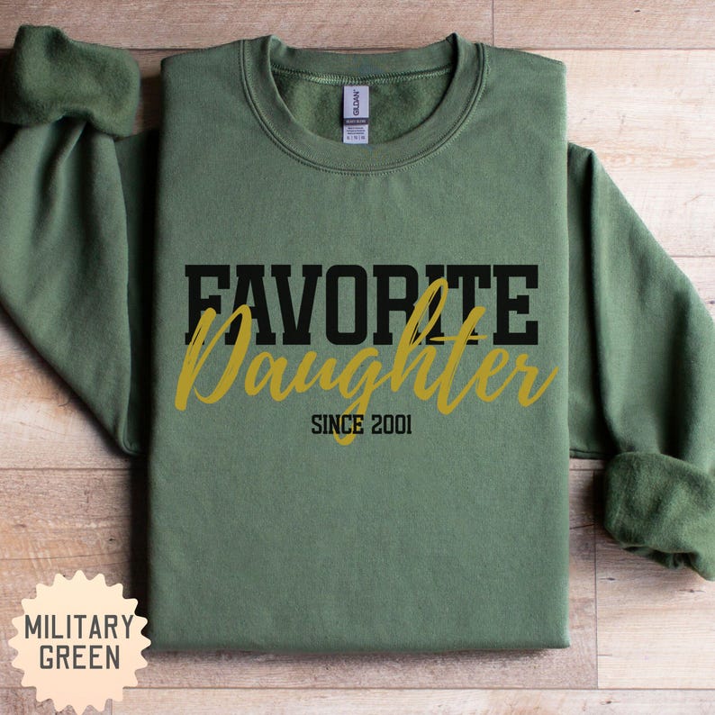 Puede incluir: Sudadera verde militar con el texto "FAVORITE Daughter SINCE 2001" en letras negras y doradas. La sudadera de cuello redondo tiene mangas largas. El color es verde militar. Una etiqueta es visible en la parte superior central.