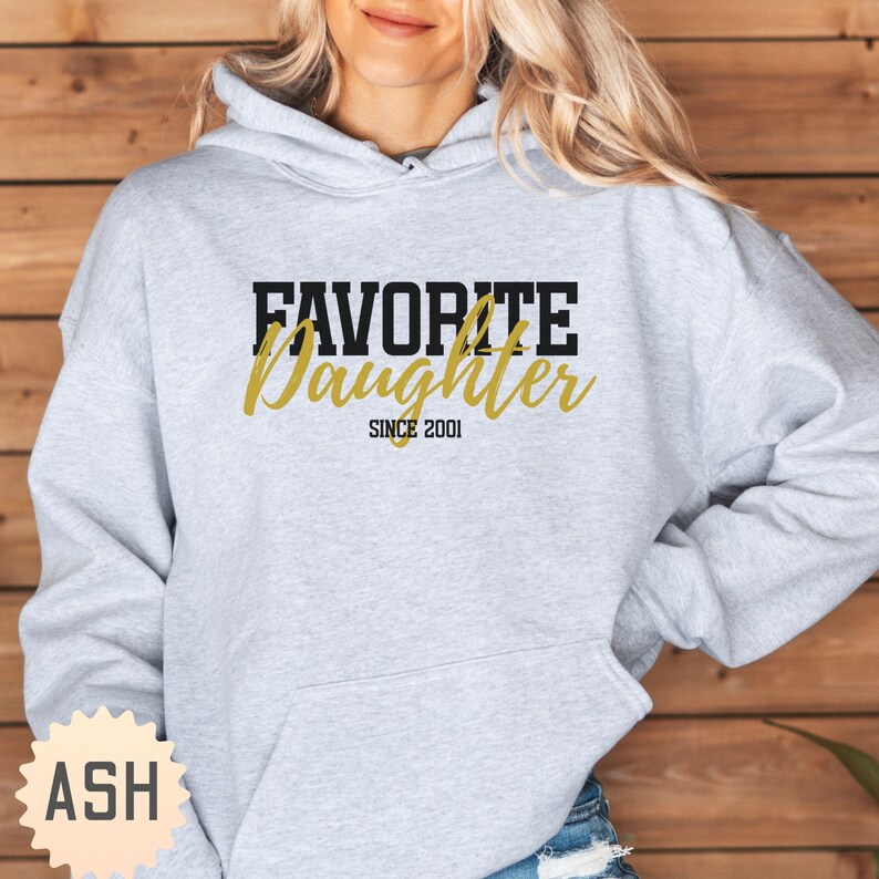 Puede incluir: Sudadera gris claro con el texto "FAVORITE Daughter SINCE 2001" en letras negras y doradas. La sudadera tiene una capucha con cord&oacute;n y un bolsillo delantero.