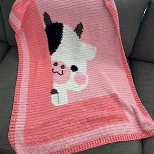 Puede incluir: Una manta de crochet rosa con un diseño de vaca blanca y negra. La manta tiene un borde rosa.