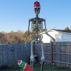 May include: A large outdoor Halloween and Christmas decoration featuring a giant skeleton wearing a Santa hat. The skeleton stands in a yard with Grinch-themed cutouts and a fence in the background. The sky is clear and blue.