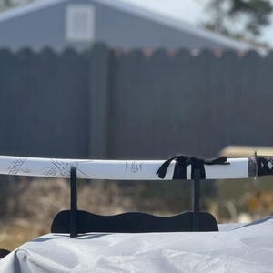 May include: A white katana sword with a black handle and a black scabbard. The sword is on a black stand and has a decorative design on the blade.