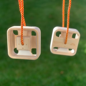 May include: Two wooden climbing holds with red and yellow rope attached. The holds are square with a hole in the center and four smaller holes around the edges. The holds are hanging against a green grassy background.