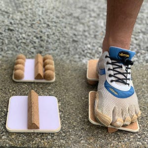 May include: A person's foot wearing a white and blue Vibram shoe steps on a small wooden platform with a cork surface. The platform is one of several similar platforms with cork surfaces, some with cork balls and others with cork triangles.