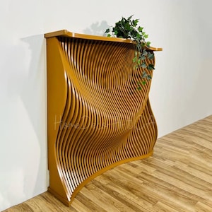 May include: A gold-coloured wooden wall panel with a wavy, three-dimensional design. The panel is made of thin strips of wood that are arranged in a curved pattern. The panel is mounted on a wall and has a plant in front of it.