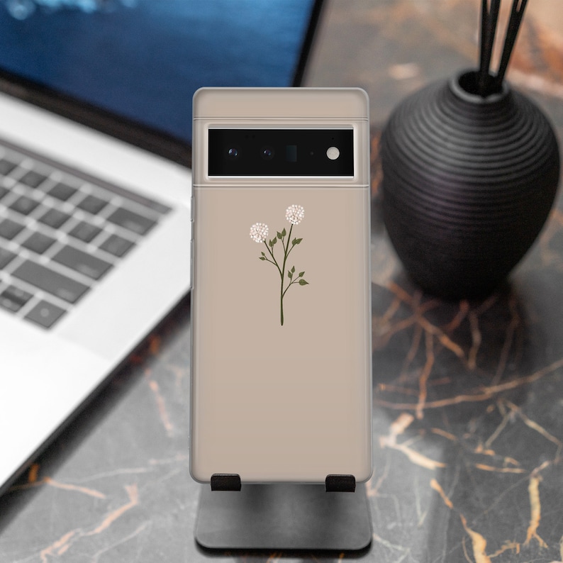 May include: A beige phone case with a simple floral design featuring two white flowers and green stems. The phone case is on a stand.