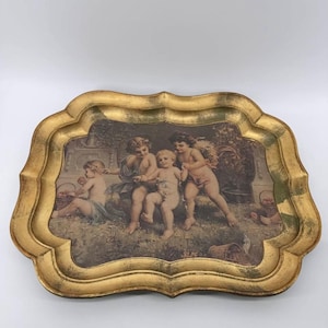 Puede incluir: Una bandeja rectangular con borde festoneado y acabado en oro envejecido. El centro presenta una ilustración impresa de cuatro querubines jugando en un jardín con cestas de frutas, en tonos tierra apagados y con suaves reflejos.