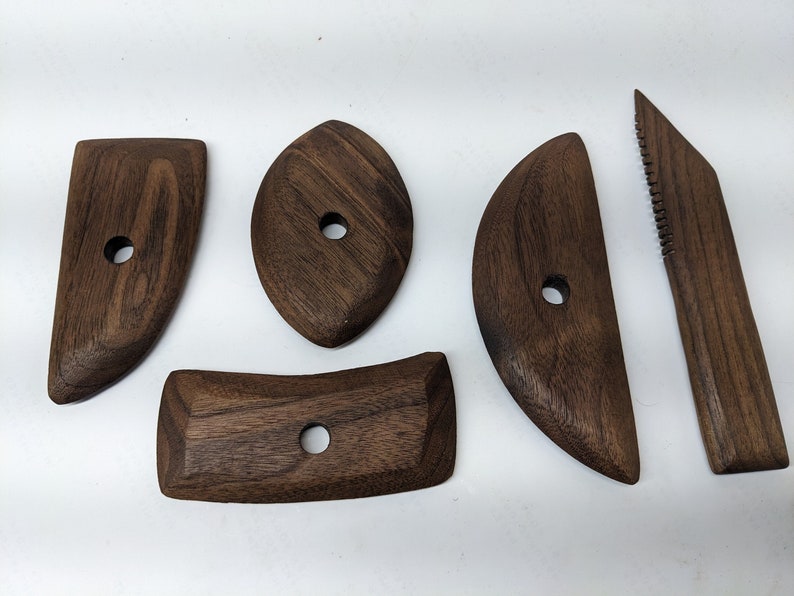 May include: Set of five wooden tools for sculpting clay. The tools are made of dark brown wood and have different shapes, including a rounded rectangle, a curved shape, and a pointed tool with a serrated edge.