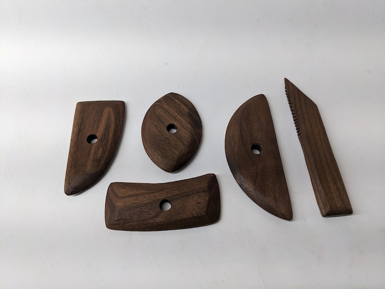 May include: Five wooden tools for sculpting clay, made of dark brown walnut wood. The tools have different shapes, including a curved blade, a rounded edge, and a flat edge.