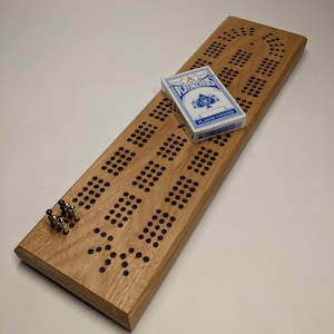 May include: A wooden cribbage board with a card on top. The board has a light brown stain and is made of solid wood. The card is a standard playing card with a blue back and the Ace of Spades face.