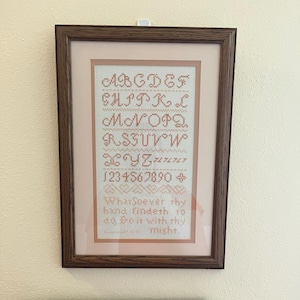 May include: A framed cross-stitch sampler featuring the alphabet, numbers, and the quote "Whatsoever thy hand findeth to do, Do it with thy might." The embroidery is in shades of peach and cream, set against a light pink background.