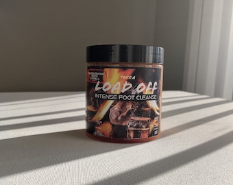 May include: A glass jar of foot scrub with a black lid and a label that says "Take a Load Off Intense Foot Cleanse". The label has a fiery orange background with a graphic of burning logs.