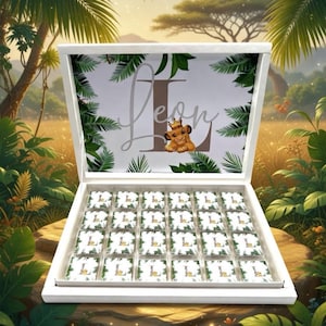 Könnte beinhalten: Eine weiße Schachtel voller Pralinen, verziert mit dem Namen "Leon" und einem Cartoon-Löwen mit Krone. Die Pralinen sind einzeln verpackt mit passendem Design, vor dem Hintergrund eines Dschungels.