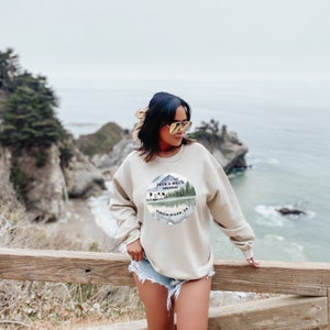 May include: A beige sweatshirt with a circular graphic of a camper van in front of a mountain and trees. The text "Jack & Mel's Airstream Virgin River, CA" is printed around the graphic.