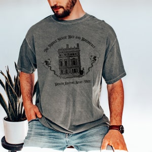 May include: Gray t-shirt with a graphic featuring a detailed illustration of a building, two skulls, and the text "THE RIDDLE HOUSE BED AND BREAKFAST" and "DEATH EATERS STAY FREE". The shirt has a vintage, worn look.