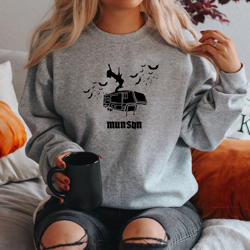 May include: A gray sweatshirt with a black graphic of a person standing on top of a camper with bats flying around. The text "munson" is printed below the camper.