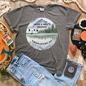May include: A dark gray t-shirt with a white circle graphic featuring a vintage Airstream trailer in a forest setting. The text "Jack & Mel's Airstream Virgin River, CA" is printed on the graphic.