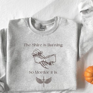 May include: A gray sweatshirt with the text "The Shire is Burning So Mordor it is" and a drawing of hands holding a book. A winged sword is below the text.