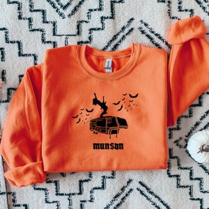 May include: Orange sweatshirt with a black graphic of a person standing on top of a camper with bats flying around. The text "munson" is printed below the camper.