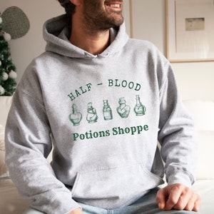 Könnte beinhalten: Ein grauer Kapuzenpullover mit dem Aufdruck "HALF - BLOOD Potions Shoppe" auf der Vorderseite. Der Text ist grün und es gibt fünf kleine Zeichnungen von Zaubertrankflaschen in grün.