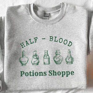 Könnte beinhalten: Grauer Pullover mit dem Text "HALF - BLOOD" und einer Reihe von fünf Zaubertrankflaschen mit dem Text "Potions Shoppe" darunter.