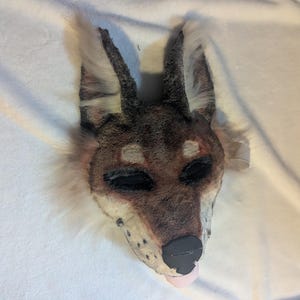 May include: A handmade dog mask with a brown and white fur design. The mask features black eye sockets, a black nose, and a pink tongue. The ears are upright and pointed, and the mask is set against a white background.