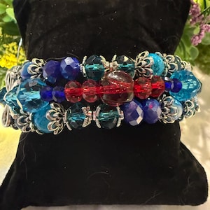 May include: A multi-strand stretch bracelet featuring faceted beads in shades of blue, teal, and red, interspersed with silver-toned metal filigree spacers and decorative end caps, arranged in a layered design on a black velvet display cushion.